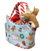 A plush brown deer toy peeking out of a light blue fabric tote bag decorated with red-and-white striped handles and Christmas-themed illustrations, set on a wooden surface.